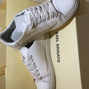 Vita sneakers från Axel Arigato - Säljer ett par stilrena vita sneakers från Axel Arigato. Skorna har en snygg broderad fågel på sidan och är i mycket bra skick. Perfekta för både vardag och fest! De har en klassisk snörning och är gjorda i skinn för en lyxig känsla.