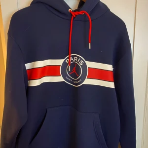Blå hoodie med Paris Saint-Germain tryck - Säljer en snygg blå hoodie från Jordan med Paris Saint-Germain tryck. Den har en röd och vit rand över bröstet och en praktisk magficka. Perfekt för kyliga dagar eller när du vill ha en sportig look. Huvan har röda snören för justering. Passar både till vardags och sportiga aktiviteter.