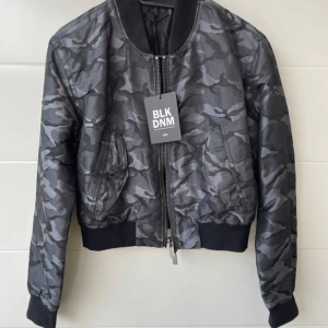 Camouflage bomberjacka från BLK DNM - Snygg kort bomberjacka i grått camouflage från BLK DNM. Jackan har en dragkedja framtill och ribbade muddar vid ärmslut och nederkant. Perfekt för en cool och avslappnad stil.