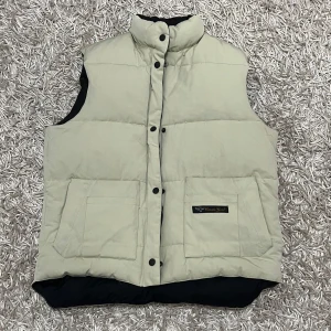 Canada Goose väst - Säljer en stilren beige dunväst från Canada Goose med svarta detaljer. Västen har en hög krage och knappar framtill. Perfekt för kyliga dagar när du vill hålla värmen utan att kompromissa med stilen. (Pris kan diskuteras)❗️