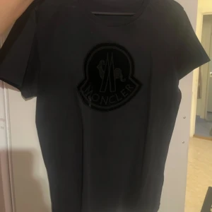 Svart t-shirt från Moncler - Snygg svart t-shirt från Moncler med ett stort, stilrent logotryck på bröstet. Perfekt för en casual look med en touch av lyx. T-shirten har en rund halsringning och korta ärmar.Använd några gånger, köpt från Wakakuu