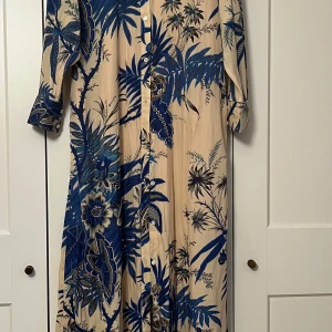 Banana Republic klänning  - Elegant långklänning i beige med blått blommönster. Klänningen har långa ärmar och knappar framtill samt ett matchande bälte i midjan. Storlek L. Nypris ca 1500kr. Nyskick. 