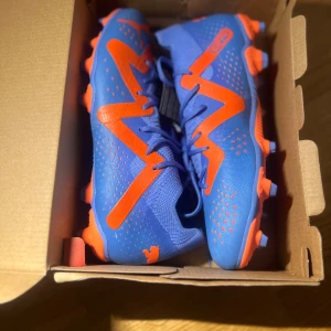 Blå och orange fotbollsskor från Puma - Säljer ett par snygga fotbollsskor från Puma i blått och orange. Skorna har en cool design med snörning och är perfekta för fotbollsträning eller match. De är i nytt skick och kommer i originalkartong. Perfekt för dig som vill sticka ut på planen! ⚽️