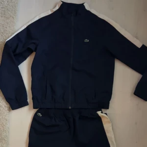 Mörkblå Lacoste set (Zip Tröja & Byxa) (nypris: 1699kr) - Säljer ett snyggt och trendigt Lacoste set köpt från kidsbrandstore för 1699kr. Zip hoodien har en dragkedja framtill och långa ärmar. Den har en stilren design med vita detaljer längs ärmarna med ”Lacoste” text och det klassiska Lacoste-märket på bröstet och byxorna.