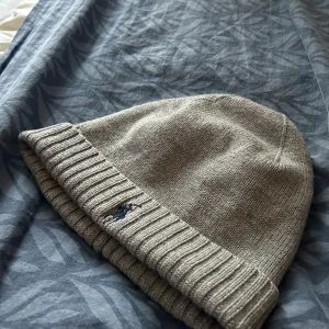 Grå stickad mössa - Säljer en snygg grå stickad mössa i mycket bra skick. Den har en ribbad kant och en liten broderad detalj på sidan. Perfekt för höst och vinter för att hålla dig varm och stilren. Passar de flesta tack vare sin flexibla passform.