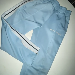 Ljusblå byxor från Palm Angels - Säljer ett par ljusblå träningsbyxor från Palm Angels i storlek M. Byxorna har en vit sidoremsa och dragkedja vid bensluten. Perfekta för en avslappnad stil eller träning. De är i mycket bra skick och redo för nya äventyr!