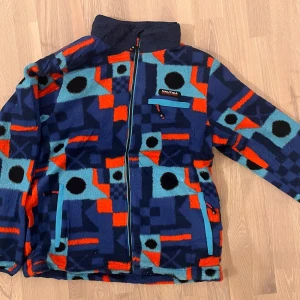Vintage Crazy Fleece - Färgglad fleecejacka från Nautica - Säljer en färgglad fleecejacka från Nautica med ett unikt geometriskt mönster i blått, rött och turkost. Jackan har en dragkedja framtill och praktiska fickor. Perfekt för att sticka ut i mängden!