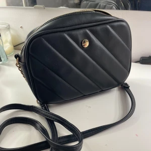 Svart quiltad axelväska - Snygg svart axelväska i quiltat mönster med dragkedja och justerbar axelrem. Perfekt för att bära dina nödvändigheter med stil.