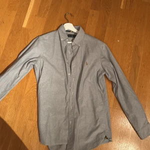 Grå skjorta från Ralph Lauren - Säljer en stilren grå skjorta från Ralph Lauren i mycket bra skick. Skjortan är långärmad och har en klassisk krage med knappar. Perfekt för både vardag och festliga tillfällen. Den är gjord i ett mjukt bomullsmaterial som känns skönt mot huden. Passar perfekt till både jeans och chinos! Size -S , inga defekter 