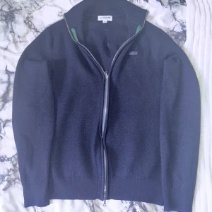 Mörkblå ullkofta från Lacoste - Säljer en snygg mörkblå kofta från Lacoste i 100% ull. Den har en dragkedja framtill och det klassiska Lacoste-märket på bröstet. Perfekt för höst och vinter när du vill hålla dig varm och stilren. Passar både till vardags och lite finare tillfällen.     Ny pris på varan närmare 2000kr mitt pris 1000kr kan gå ner vid snabb affär endast!
