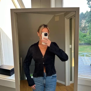 Svart blus med knappar - Säljer en snygg svart blus med knappar framtill. Den har en v-ringad design och långa ärmar, perfekt för både vardag och fest. Materialet ser ut att vara lätt och bekvämt, vilket gör den idealisk för olika tillfällen.