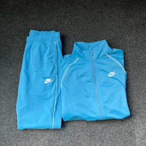 Blå träningsset från Nike - Säljer ett snyggt blått träningsset från Nike. Setet består av en långärmad jacka med dragkedja och matchande byxor. Perfekt för träning eller en avslappnad dag. Nike-loggan syns tydligt på både jackan och byxorna. Setet är i bra skick och redo för nya äventyr!