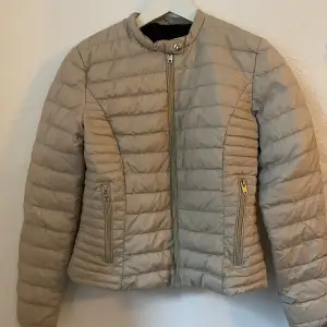 Säljer en snygg beige pufferjacka från Rockandblue i storlek 36. Jackan är quiltad och har en dragkedja framtill samt två dragkedjeförsedda fickor. Den är tillverkad i polyester och är perfekt för höst och vår. Jackan är i bra skick och redo för nya äventyr!