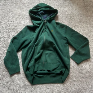 Grön hoodie från Polo Ralph Lauren - Skön slutsåld zip hoodie från Polo Ralph Lauren i praktiskt taget nyskick. Den är mörkgrön med matchande blå-grönt rutigt mönster i huvan. Broderad gul logga på bröstet. Hatar att sälja den men har vuxit ur den. 