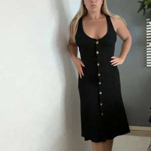 Svart ribbad klänning från ONLY - Svart ribbad klänning från ONLY. Tight, klassisk passform och knappar framtill som ger en stilren look. Klänningen är ärmlös och har en v-ringad urringning, perfekt för både vardag och fest. Materialet är mjukt och stretchigt, vilket gör den superbekväm att bära.