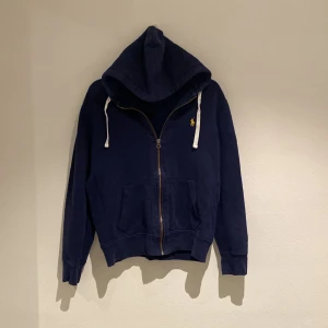 Mörkblå hoodie från Polo Ralph Lauren - Säljer en snygg och bekväm mörkblå hoodie från Polo Ralph Lauren. Den har en dragkedja framtill och en klassisk huva med vita snören. Perfekt för både höst och vinter, och den är i bra skick. Passar perfekt för en avslappnad stil!