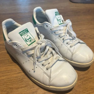 Vita Adidas Stan Smith sneakers - Säljer ett par klassiska vita Adidas Stan Smith sneakers med gröna detaljer. Skorna har snörning och är i skinn. De är lite slitna men fortfarande i bra skick. Perfekta för både vardag och fest!