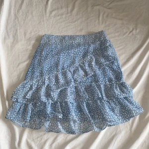 Blå blommig kjol med volanger - Säljer en superfin blå kjol med ett sött blommönster. Kjolen har flera lager av volanger som ger den en härlig och lekfull look. Perfekt för sommardagar eller en festlig tillställning. Den är i bra skick och redo för nya äventyr!