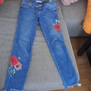 Säljer ett par snygga blå jeans med blommigt broderi på benen. De har en skinny passform och är i ett välanvänt skick. Perfekta för en casual look med en twist! De har en klassisk femficksdesign och en dragkedja med knapp framtill.