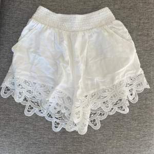 Säljer ett par supercharmiga vita shorts med bred resår i midjan för en bekväm passform. De har vackra spetsdetaljer längs nederkanten som ger en romantisk touch. Perfekta för sommardagar eller en avslappnad fest! 🕊️