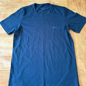 Mörkblå t-shirt från Boss - Säljer en snygg mörkblå t-shirt från Boss i storlek M. Den är kortärmad och har en stilren design med en broderad Boss-logga på bröstet. Perfekt för både vardag och fest!