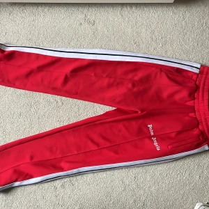 Palm Angles byxor - Snygga röda träningsbyxor från Palm Angels med vita ränder längs sidorna. Byxorna har en elastisk midja och är perfekta för både träning och avslappnade dagar. Märkesloggan är tryckt i vitt på ena benet. Superbekväma och stilrena!