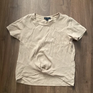 Beige ribbad topp från XOXO Friends - Säljer en superfin beige ribbad topp från XOXO Friends. Den har korta ärmar och en snygg knutdetalj framtill. Perfekt för en avslappnad vardagslook eller att styla upp med accessoarer. Toppen är i bra skick och redo för en ny ägare!