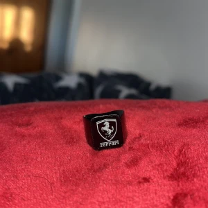 Svart ring med Ferrari-logga - En Cool svart ring med den ikoniska Ferrari-loggan i silver. Perfekt för bilentusiaster eller som en statement-accessoar. Ringen har en blank yta och känns både modern och stilren. Passar till både vardag och fest!
