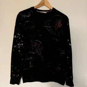 Svart tröja från Moncler - Säljer en svart tröja från Moncler i bra skick. Den har ett unikt stänkmönster i vitt och ett broderat Moncler-märke på ärmen. Perfekt för höst och vinter, med långa ärmar. Den är storlek M men den kan även passa S