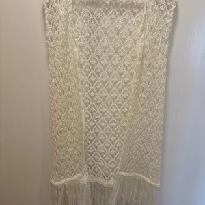 Vit spetskofta från Elitza - Säljer en vacker vit spetskofta från Elitza i storlek M. Klänningen har ett elegant mönster och fransar längst ner som ger en bohemisk känsla. Perfekt för sommarens fester eller en dag på stranden. Den är ärmlös och har en luftig passform.