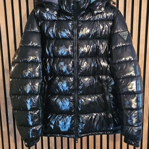 Svart glansig pufferjacka från Guess - Säljer en svart glansig pufferjacka från Guess i mycket bra skick. Jackan har en cool, quiltad design med dragkedja och en galaktisk mönstrad insida. Perfekt för vintern med sin varma och stiliga look. Passar både till vardags och festliga tillfällen!