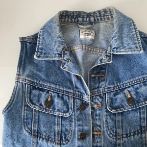 Blå jeansväst från Chic Avenue - Säljer en snygg blå jeansväst från Chic Avenue i storlek M. Västen har en klassisk krage och knappar framtill. Två bröstfickor med knappdetaljer ger en cool look. Perfekt för vår och sommar!