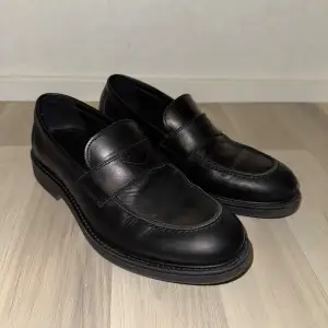 Snygga svarta loafers i skinn, perfekta för både vardag och fest. De har en klassisk design med en bred rem över foten och en robust sula. Skorna är i mycket bra skick och passar perfekt till både jeans och kostym. Ett måste i garderoben för den stilmedvetne!