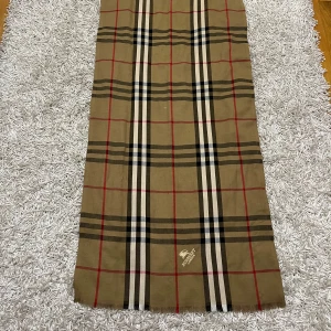 Burberry halsduk - Säljer en klassisk rutig halsduk från Burberry i beige, svart, vit och röd. Den är perfekt för att hålla dig varm och stilren under höst och vinter. Halsduken är i mycket bra skick och har den ikoniska Burberry-designen. Perfekt accessoar för både vardag och fest! (Priset kan diskuteras)