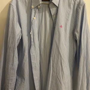 Blåvit randig skjorta från Morris - Säljer en stilren blåvit randig skjorta från Morris i storlek S. Skjortan har långa ärmar och knappar framtill. Perfekt för både vardag och festliga tillfällen. Den är i mycket bra skick och har en liten rosa broderad detalj på bröstet. Passar perfekt till både jeans och kostymbyxor!