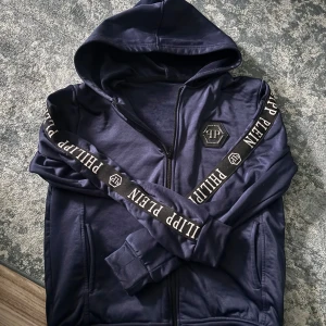 Mörkblå hoodie från Philipp Plein - Säljer en snygg mörkblå hoodie från Philipp Plein med dragkedja och huva. Den har långa ärmar med märkets namn tryckt längs ärmarna. Perfekt för en casual look eller att mysa i hemma. Passar både till vardags och chill.