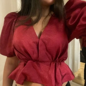 Röd topp med puffärmar - Säljer en superfin röd topp med puffiga kortärmar och volangdetalj i midjan. Toppen har en djup v-ringning och är perfekt för festliga tillfällen. Materialet ser ut att vara glansigt och ger en lyxig känsla. Passar perfekt till både kjol och byxor! Prislapp sitter också kvar💞