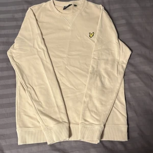 Beige tröja från Lyle & Scott - Säljer en stilren beige tröja från Lyle & Scott i mycket bra skick. Den har en broderad gul logga på bröstet och är perfekt för både höst och vår. Tröjan är långärmad och gjord i mjukt material för extra komfort. Passar perfekt till vardags eller en avslappnad dag ute. Pris kan diskuteras!