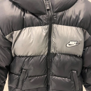 Svart och grå pufferjacka från Nike - Säljer en snygg och varm pufferjacka från Nike i svart och grått. Jackan har en dragkedja framtill och är perfekt för kalla dagar. Den är i mycket bra skick och har en Therma-Fit teknologi för extra värme. Passar perfekt för vintern! Jackan är för barn! 