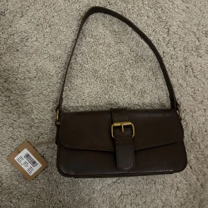 Brun axelväska brandy melville (nypris 400kr) - Snygg brun axelväska i fakeskinn med guld spänne. Stilren till olika outfits. Väskan är i nytt skick, helt oanvänd från brandy melville. Säljer då den ej passade min stil 💕 