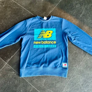 Blå sweatshirt från New Balance - Säljer en snygg blå sweatshirt från New Balance. Den har ett stort tryck med loggan i gult och blått på framsidan. Perfekt för en avslappnad stil eller träning. Tröjan är i bra skick och har långa ärmar. Passar perfekt till både vardag och sportiga aktiviteter.