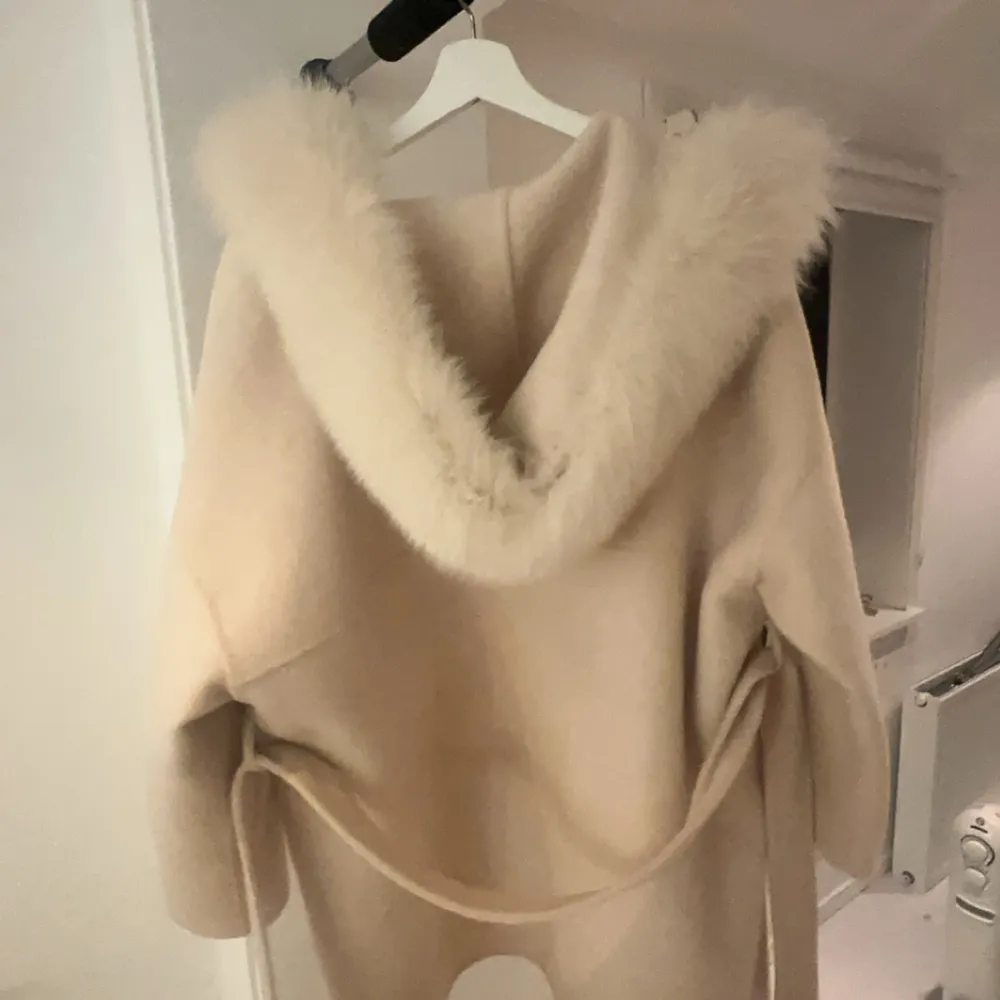 Säljer en superfin beige kappa med fluffig pälshuva. Den är perfekt för vintern och har ett bälte i midjan för en snygg passform. Kappan har stora fickor och är långärmad, vilket gör den både praktisk och stilren. Perfekt för både vardag och fest!. Jackor.