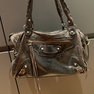 Baleciaga liknande handväska - Säljer en cool svart handväska i skinn med dragkedja och fransdetaljer. Väskan har en sliten look som ger den en unik stil. Perfekt för vardagsbruk eller en utekväll. Inuti finns ett leopardmönstrat foder som ger en extra touch av stil. Väskan är välanvänd och behöver piffas upp lite. 