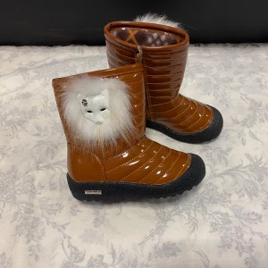 Bruna vinterstövlar med pälsdetalj - Säljer ett par unika bruna vinterstövlar med en cool pälsdetalj och maskmotiv. De har en glansig yta och dragkedja på sidan för enkel påtagning. Insidan är fodrad för extra värme. Perfekta för kalla vinterdagar!                          Stlk kvar  27,29     Med fleece inuti, rekommenderar att välja en Stlk större 