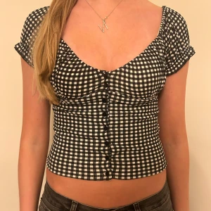 Svartvit rutig blus - Snygg och trendig svartvit rutig blus med korta ärmar och knappar framtill. Den har en v-ringad design och är croppad, vilket gör den perfekt för sommardagar. Passar både till vardags och festliga tillfällen.
