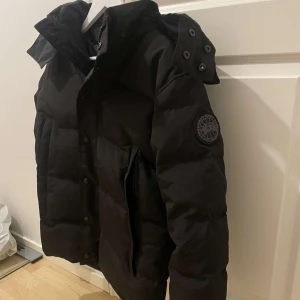 Svart dunjacka från Canada Goose - Säljer en svart dunjacka från Canada Goose i mycket bra skick. Jackan har en avtagbar huva och stängs med både dragkedja och knappar. Perfekt för kalla vinterdagar med sin varma och isolerande design. Den har långa ärmar och ribbstickade muddar för extra komfort.