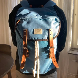 Blå ryggsäck från Sandqvist - Säljer en snygg blå ryggsäck från Sandqvist. Den har bruna läderdetaljer och en praktisk snörning. Perfekt för vardagsbruk eller utflykter. Ryggsäcken är i mycket bra skick och redo för nya äventyr!