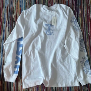 Vit långärmad tröja från Vailent - Säljer en snygg vit långärmad tröja från Vailent med blått tryck på bröstet och ärmen. Perfekt för en avslappnad stil och passar bra till både jeans och shorts. Tröjan är i nytt skick med etikett kvar. Perfekt för vår och sommar! Nypris 499kr