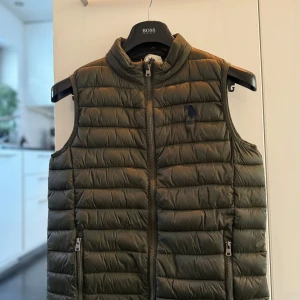Grön  väst - Säljer en snygg grön dunväst från USPA i mycket bra skick. Storlek 14-15 men passar small. Västen har en dragkedja framtill och är quiltad för extra värme. Perfekt för höst och vår, med en broderad logga på bröstet. Passar både till vardags och lite finare tillfällen. Frakt ingår. 