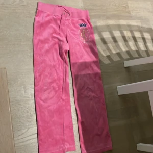 Rosa mjukisbyxor från Juicy Couture - Supermysiga rosa mjukisbyxor från Juicy Couture med en glittrig logga och krona på sidan. De har en bekväm resårmidja med snörning för perfekt passform. Perfekta för en chill dag hemma eller en avslappnad utflykt. 🩷